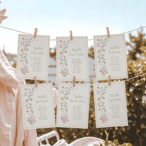 May include: Six rectangular table cards with floral designs and the words "Table Thirteen" through "Table Eighteen" are displayed. Each card is held by a wooden clothespin on a string. The cards have a light green border and a floral design.