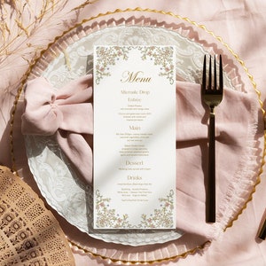 May include: A white wedding menu card with gold floral accents and the word "Menu" in cursive. The card is placed on a white plate with a pink napkin and gold cutlery.