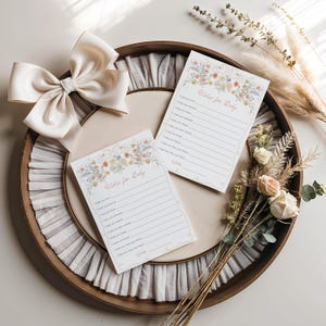 Pu&ograve; includere: Un vassoio di legno contiene due biglietti "Wishes for Baby" con bordi floreali e spazi a righe per la scrittura. Un grande fiocco color crema e un bouquet di fiori ed erbe secche completano la scena, creando una palette di colori morbida e neutra.