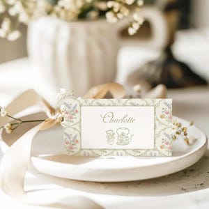 May include: A white place card with a green floral border and the name "Charlotte" printed in gold script. A small teapot and teacup are illustrated in the bottom right corner.