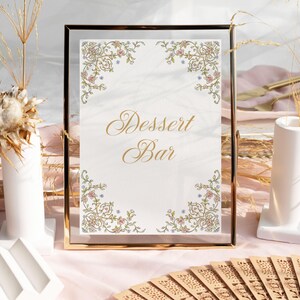 Op de afbeelding: Een gouden lijst met een witte achtergrond en gouden letters die "Dessert Bar" lezen. Het bord is versierd met een bloemmotief in roze, blauwe en groene tinten. Het bord staat op een tafel met een roze tafelkleed.