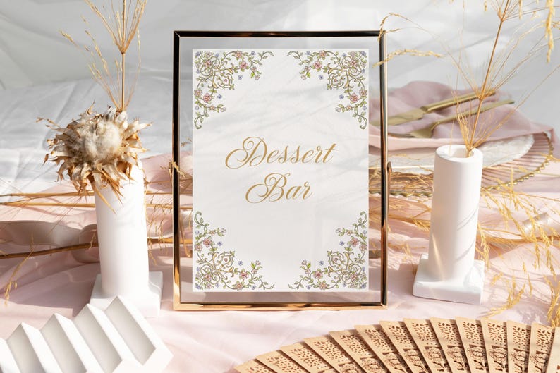 Op de afbeelding: Een gouden lijst met een witte achtergrond en gouden letters die "Dessert Bar" lezen. Het bord is versierd met een bloemmotief in roze, blauwe en groene tinten. Het bord staat op een tafel met een roze tafelkleed.
