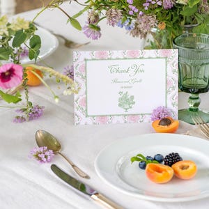 May include: A rectangular thank you card with a pink floral border and green accents, featuring the text Thank You, names, a date, and a green botanical illustration. The card is displayed on a table with a white plate, fruit, and glassware.
