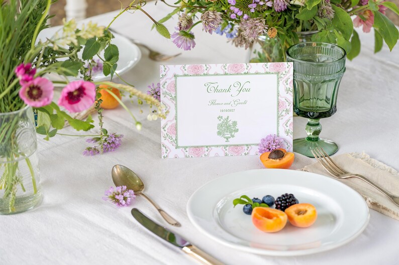 May include: A rectangular thank you card with a pink floral border and green accents, featuring the text Thank You, names, a date, and a green botanical illustration. The card is displayed on a table with a white plate, fruit, and glassware.