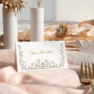 May include: A white place card with a floral border and the text "Your Text Here" in gold lettering. The place card is on a pink tablecloth with a gold fork.
