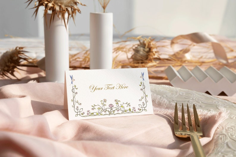 May include: A white place card with a floral border and the text "Your Text Here" in gold lettering. The place card is on a pink tablecloth with a gold fork.