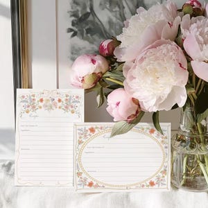 May include: Two blank recipe cards with floral borders. The cards are white with a gold border and a floral design. The cards are on a white surface with a bouquet of pink peonies in the background.