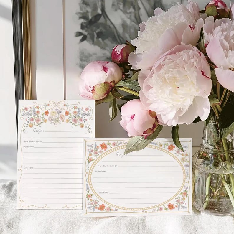 May include: Two blank recipe cards with floral borders. The cards are white with a gold border and a floral design. The cards are on a white surface with a bouquet of pink peonies in the background.