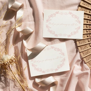May include: Two white cards with pink floral borders and the text "Thank you for showering us with love" on a pink fabric background with a wooden fan and dried flowers.