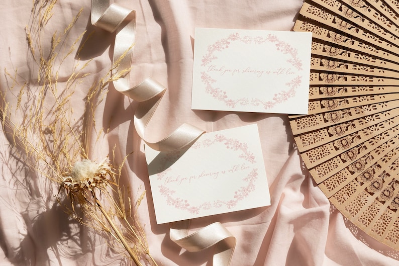May include: Two white cards with pink floral borders and the text "Thank you for showering us with love" on a pink fabric background with a wooden fan and dried flowers.