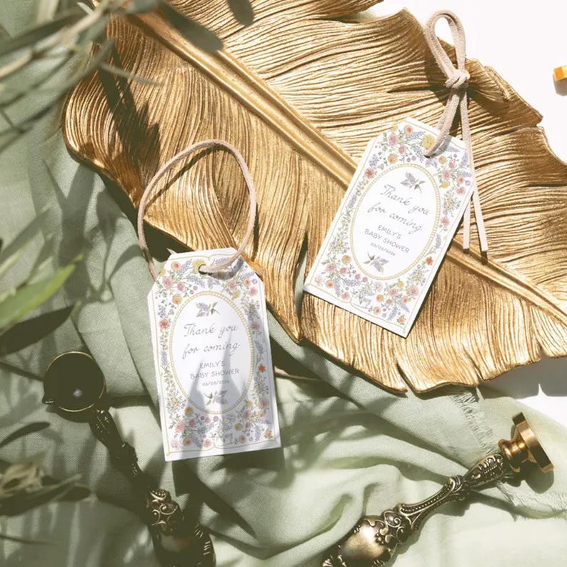 May include: Two floral baby shower gift tags with the text "Thank you for coming" and the name "Emily's Baby Shower". The tags are tied with a beige string and are on a gold leaf-shaped tray.