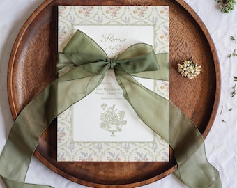 May include: A wedding invitation with floral design and the text "Florence & Co." is tied with a sheer olive green ribbon. The invitation rests on a round wooden tray, with small floral accents scattered around.