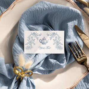 May include: A white place setting with a blue napkin, gold silverware, and a white card with a blue teapot illustration and the text "Your Text".