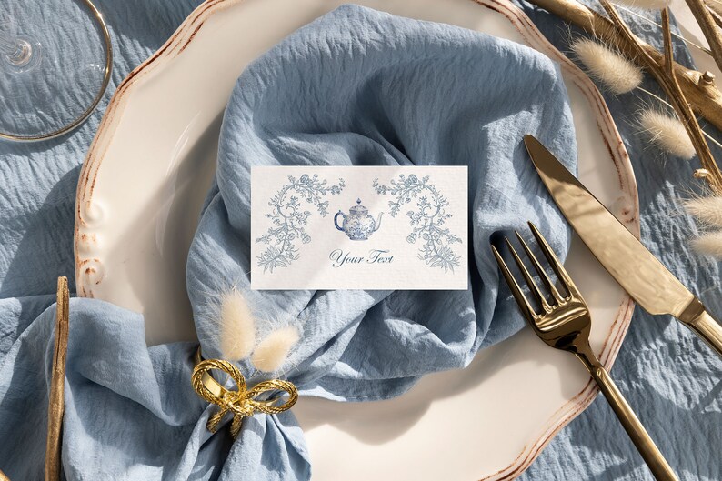 May include: A white place setting with a blue napkin, gold silverware, and a white card with a blue teapot illustration and the text "Your Text".