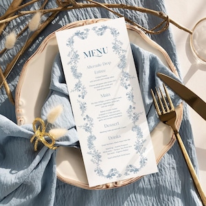 May include: A menu card with a floral border and text, placed on a white plate with a blue napkin. A gold fork and knife are on the right. A gold bow napkin ring and dried flowers are on the left. The menu is for an alternate drop.