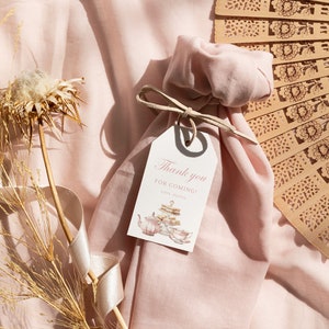 May include: A pink fabric bag with a white tag that says "Thank you for coming!" with a teacup and teapot illustration. The bag is tied with a brown ribbon and is sitting on a pink fabric surface with a brown wooden fan.