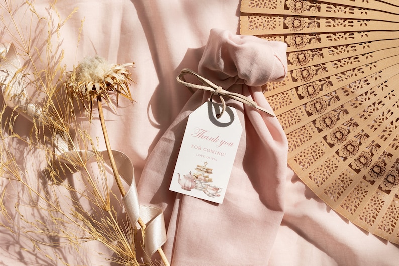 May include: A pink fabric bag with a white tag that says "Thank you for coming!" with a teacup and teapot illustration. The bag is tied with a brown ribbon and is sitting on a pink fabric surface with a brown wooden fan.