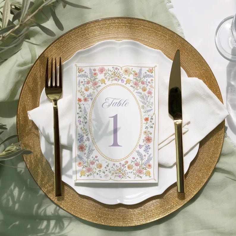 K&ouml;nnte beinhalten: Ein Gedeck mit einem goldenen Platzteller, einem wei&szlig;en Teller und einer floralen Tischnummerkarte. Die Karte zeigt das Wort &bdquo;Table&ldquo; und die Nummer &bdquo;1&ldquo;. Eine goldene Gabel und ein Messer befinden sich auf beiden Seiten der Karte.