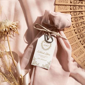 Op de afbeelding: Een roze stoffen tas met een wit label waarop 'Bedankt' staat en een gouden hartontwerp. De tas is vastgebonden met een beige lint en ligt op een roze stoffen oppervlak. Op de achtergrond is een houten waaier met een bloemenpatroon te zien.