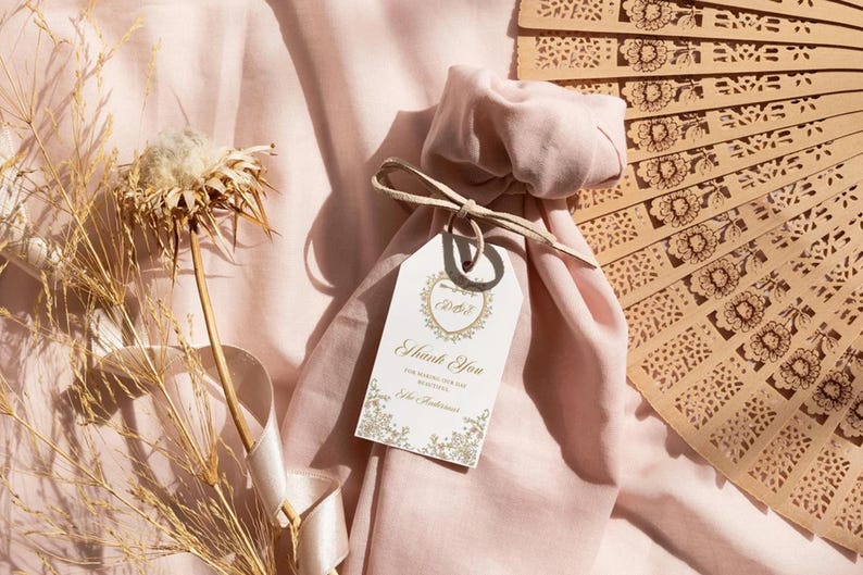 Op de afbeelding: Een roze stoffen tas met een wit label waarop 'Bedankt' staat en een gouden hartontwerp. De tas is vastgebonden met een beige lint en ligt op een roze stoffen oppervlak. Op de achtergrond is een houten waaier met een bloemenpatroon te zien.