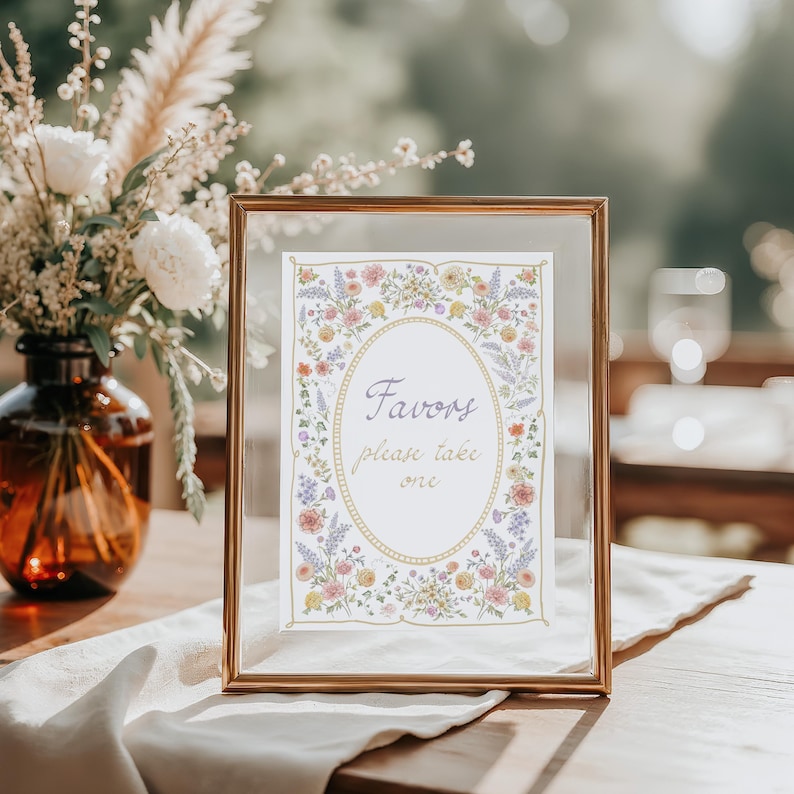 May include: A framed sign with the text "Favors please take one" in an oval shape, surrounded by floral illustrations. The frame is gold, and the sign is on a wooden table with a floral arrangement in the background.