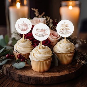 May include: Three cupcakes with white frosting topped with round paper circles featuring watercolor illustrations of a teapot, a teacup and saucer, and a tiered cake stand with sandwiches.