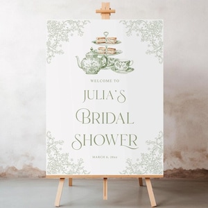 K&ouml;nnte beinhalten: Ein wei&szlig;es Schild auf einer Holzstaffelei mit dem Text "Welcome to Julia's Bridal Shower" in gr&uuml;ner Schrift. Das Schild zeigt eine gr&uuml;ne Illustration einer Teekanne, einer Teetasse und einer mehrst&ouml;ckigen Dessertplatte. Dekorative florale Elemente rahmen das obere und untere Ende ein.
