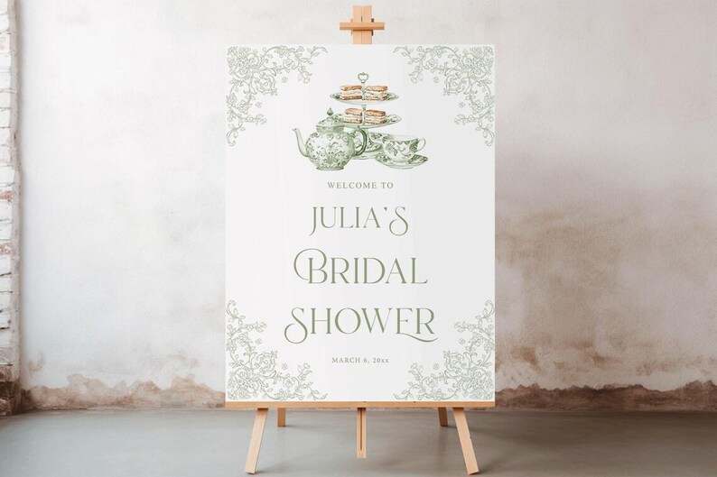 K&ouml;nnte beinhalten: Ein wei&szlig;es Schild auf einer Holzstaffelei mit dem Text "Welcome to Julia's Bridal Shower" in gr&uuml;ner Schrift. Das Schild zeigt eine gr&uuml;ne Illustration einer Teekanne, einer Teetasse und einer mehrst&ouml;ckigen Dessertplatte. Dekorative florale Elemente rahmen das obere und untere Ende ein.
