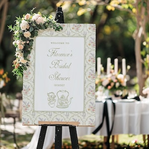 May include: A white sign with a floral pattern border, announcing "Welcome to Florence's Bridal Shower" with the date "10/10/2027". The sign is on an easel in a garden setting.
