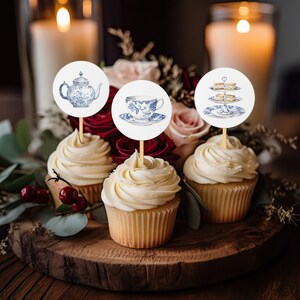 May include: Three cupcakes with white frosting topped with round paper circles featuring blue and white teacups, teapot, and a tiered cake stand. The cupcakes are on a wooden surface with a bouquet of pink and red flowers in the background.