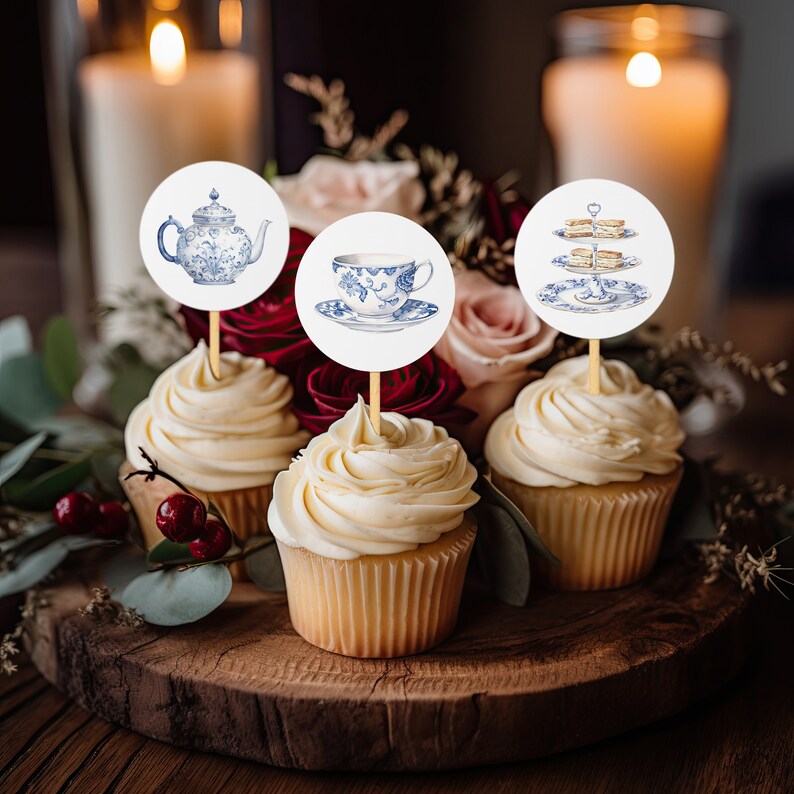 May include: Three cupcakes with white frosting topped with round paper circles featuring blue and white teacups, teapot, and a tiered cake stand. The cupcakes are on a wooden surface with a bouquet of pink and red flowers in the background.