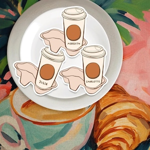 May include: Three white coffee cups with brown lids and brown coffee inside. The cups are on a white plate with the names Meredith, Julia, and Charlotte written on them. The plate is on a pink and green background with a croissant.