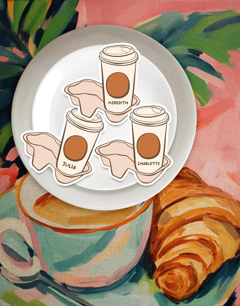 May include: Three white coffee cups with brown lids and brown coffee inside. The cups are on a white plate with the names Meredith, Julia, and Charlotte written on them. The plate is on a pink and green background with a croissant.