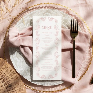 May include: A white rectangular menu card with pink floral borders and the text MENU, Alternate Drop, Entree, Main, Dessert, and Drinks. The menu is placed on a pink fabric napkin and a clear glass plate with a gold rim, alongside a black fork and knife.