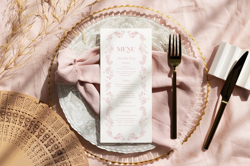 May include: A white rectangular menu card with pink floral borders and the text MENU, Alternate Drop, Entree, Main, Dessert, and Drinks. The menu is placed on a pink fabric napkin and a clear glass plate with a gold rim, alongside a black fork and knife.