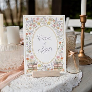 May include: A rectangular sign with the text Cards & Gifts in a script font, framed by a gold oval border and watercolor illustrations of colorful wildflowers and stacked books, held upright by a light-colored wooden block stand.