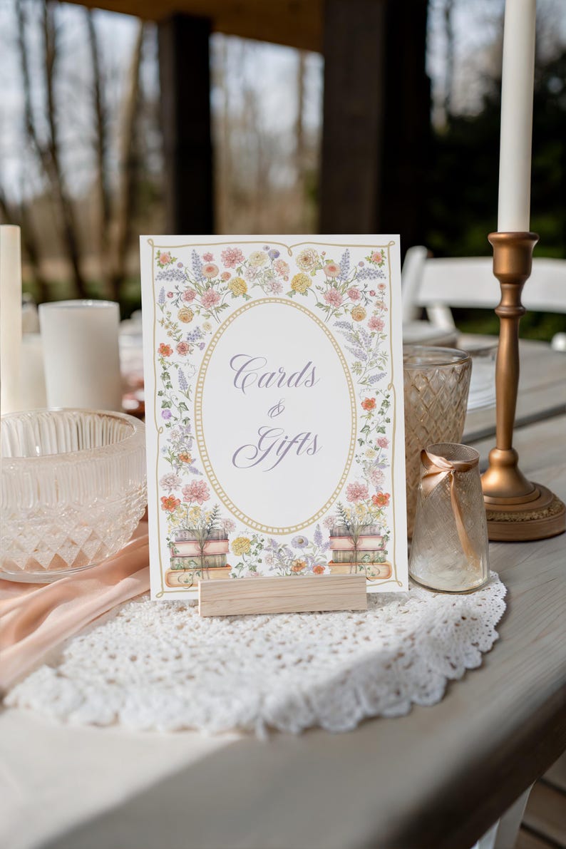 May include: A rectangular sign with the text Cards & Gifts in a script font, framed by a gold oval border and watercolor illustrations of colorful wildflowers and stacked books, held upright by a light-colored wooden block stand.