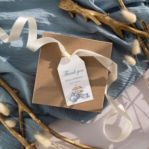 May include: A brown gift box with a white ribbon and a white tag that says "Thank you for coming! Love, The [Name]" with a blue and white illustration of a teapot and teacups. The box is on a blue fabric background with gold branches and white fluffy flowers.