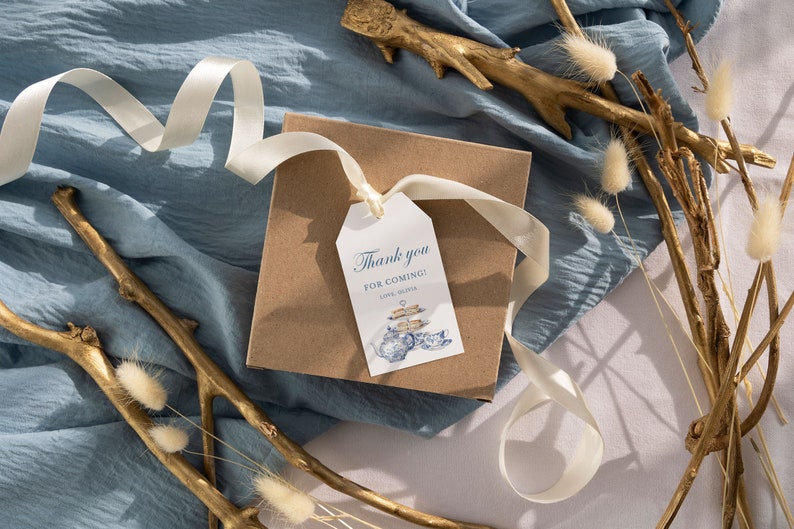 May include: A brown gift box with a white ribbon and a white tag that says "Thank you for coming! Love, The [Name]" with a blue and white illustration of a teapot and teacups. The box is on a blue fabric background with gold branches and white fluffy flowers.
