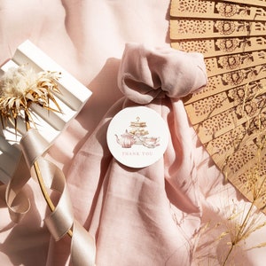 May include: A pink fabric gift bag with a round thank you tag featuring a watercolor illustration of a teapot and teacups. The tag is tied with a white ribbon. The bag is surrounded by a white ribbon, a white decorative object, and a brown wooden fan.