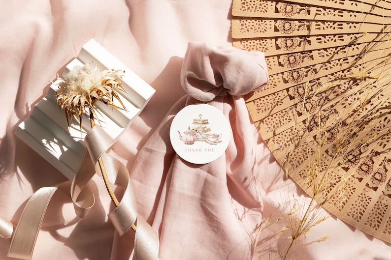 May include: A pink fabric gift bag with a round thank you tag featuring a watercolor illustration of a teapot and teacups. The tag is tied with a white ribbon. The bag is surrounded by a white ribbon, a white decorative object, and a brown wooden fan.