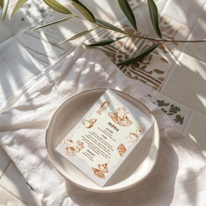 May include: A menu card with a light beige background and brown text. The menu features a latte, a pain au chocolat, and a dessert. The card is on a white plate with a light brown background.