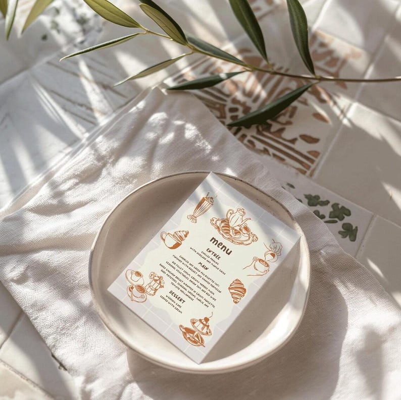 May include: A menu card with a light beige background and brown text. The menu features a latte, a pain au chocolat, and a dessert. The card is on a white plate with a light brown background.