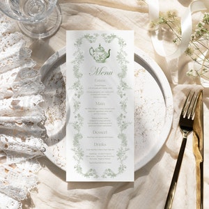 K&ouml;nnte beinhalten: Eine Men&uuml;karte mit floralem Design und dem Wort "Menu" oben. Das Men&uuml; ist von einem floralen Rand umgeben und zeigt eine Teekannen-Illustration. Ein wei&szlig;er Spitzenf&auml;cher, ein Glas und goldenes Besteck sind ebenfalls zu sehen.