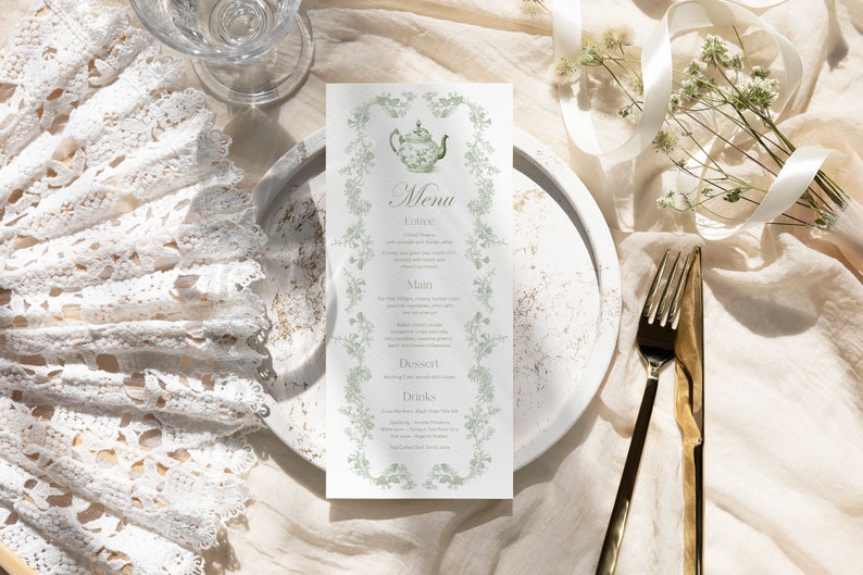 K&ouml;nnte beinhalten: Eine Men&uuml;karte mit floralem Design und dem Wort "Menu" oben. Das Men&uuml; ist von einem floralen Rand umgeben und zeigt eine Teekannen-Illustration. Ein wei&szlig;er Spitzenf&auml;cher, ein Glas und goldenes Besteck sind ebenfalls zu sehen.