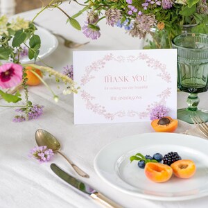 May include: A white rectangular thank you card featuring a light pink floral border, the text "THANK YOU for making our day beautiful" and "THE ANDERSONS" in pink lettering, displayed on a table with a white plate, silverware, and flowers.