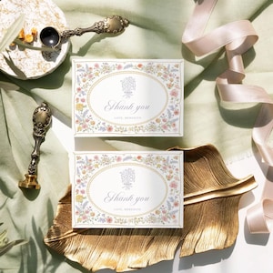 May include: Two white thank you cards with a floral border and a gold oval in the center. The text "Thank you" is written in cursive inside the oval. The top card says "Love, Meredith" and the bottom card says "Give, Meredith".