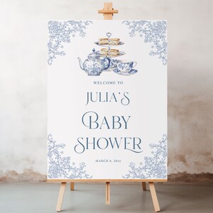 May include: A white sign with blue floral borders and a blue and white teapot, teacup, and tiered cake stand with a welcome to Julia's Baby Shower sign and the date March 6, 20xx.