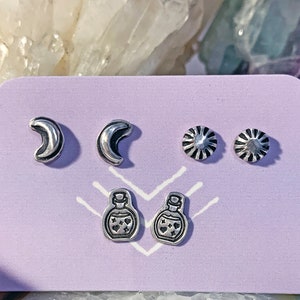May include: A set of five silver earrings on a purple card. The earrings are in the shape of moons, stars, and potion bottles. The card has the text "MahaKashi" on the bottom.