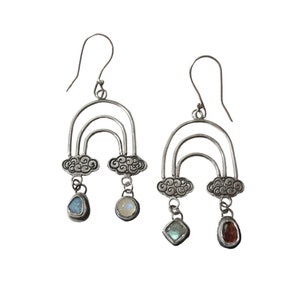 May include: Silver earrings with a rainbow and cloud design. Each earring features a teardrop-shaped gemstone and a round gemstone.